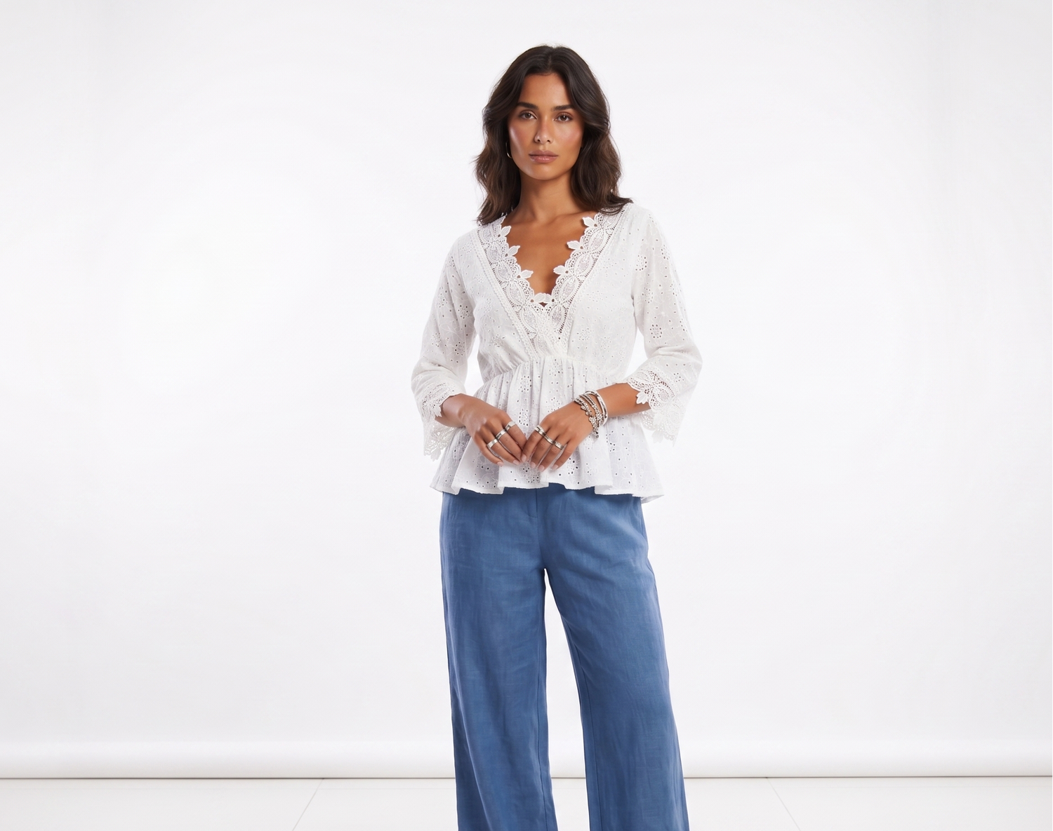 A woman modeling a white lace-trimmed long-sleeve blouse with a ruffled peplum hem, paired with blue wide-leg pants, against a plain white studio background.