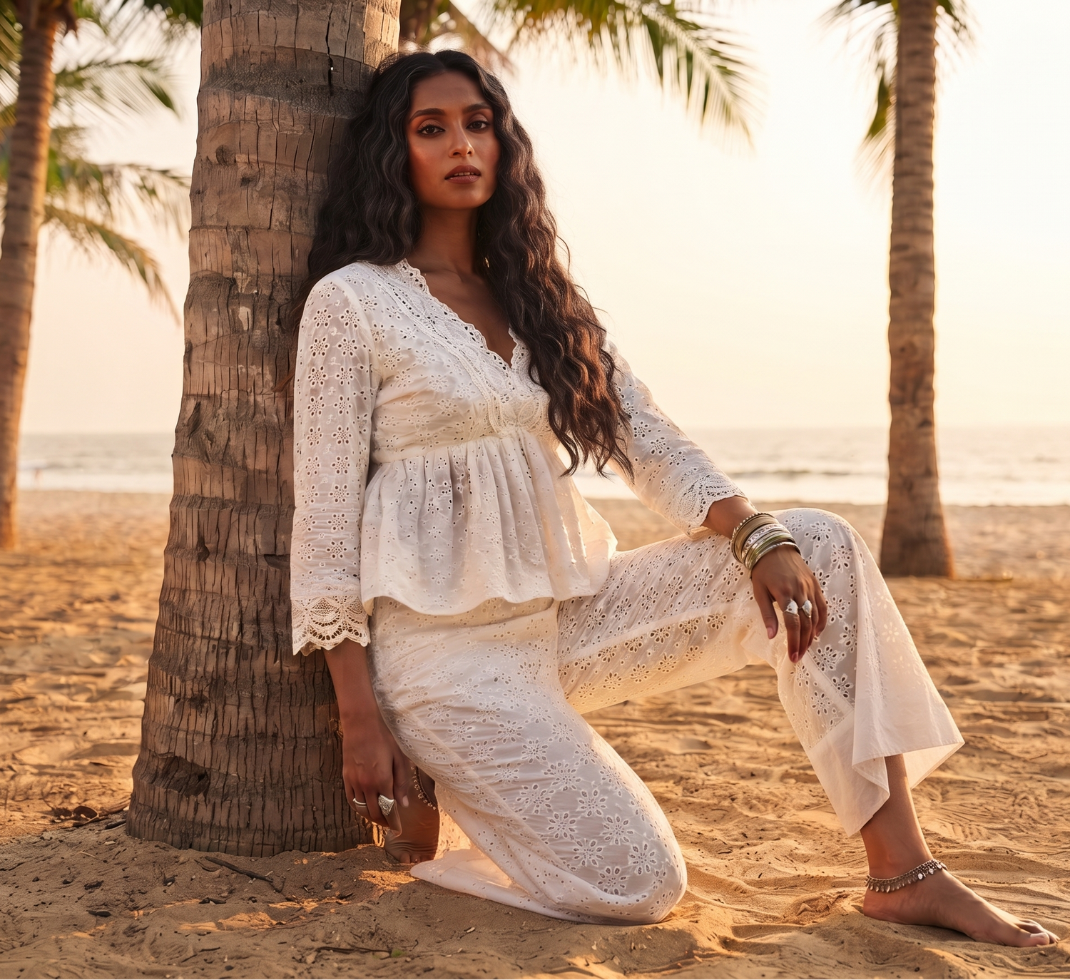 A woman kneels on a sandy beach next to a palm tree, wearing a white eyelet lace two-piece set with a peplum top and wide-leg pants. She is accessorized with multiple bracelets and rings.