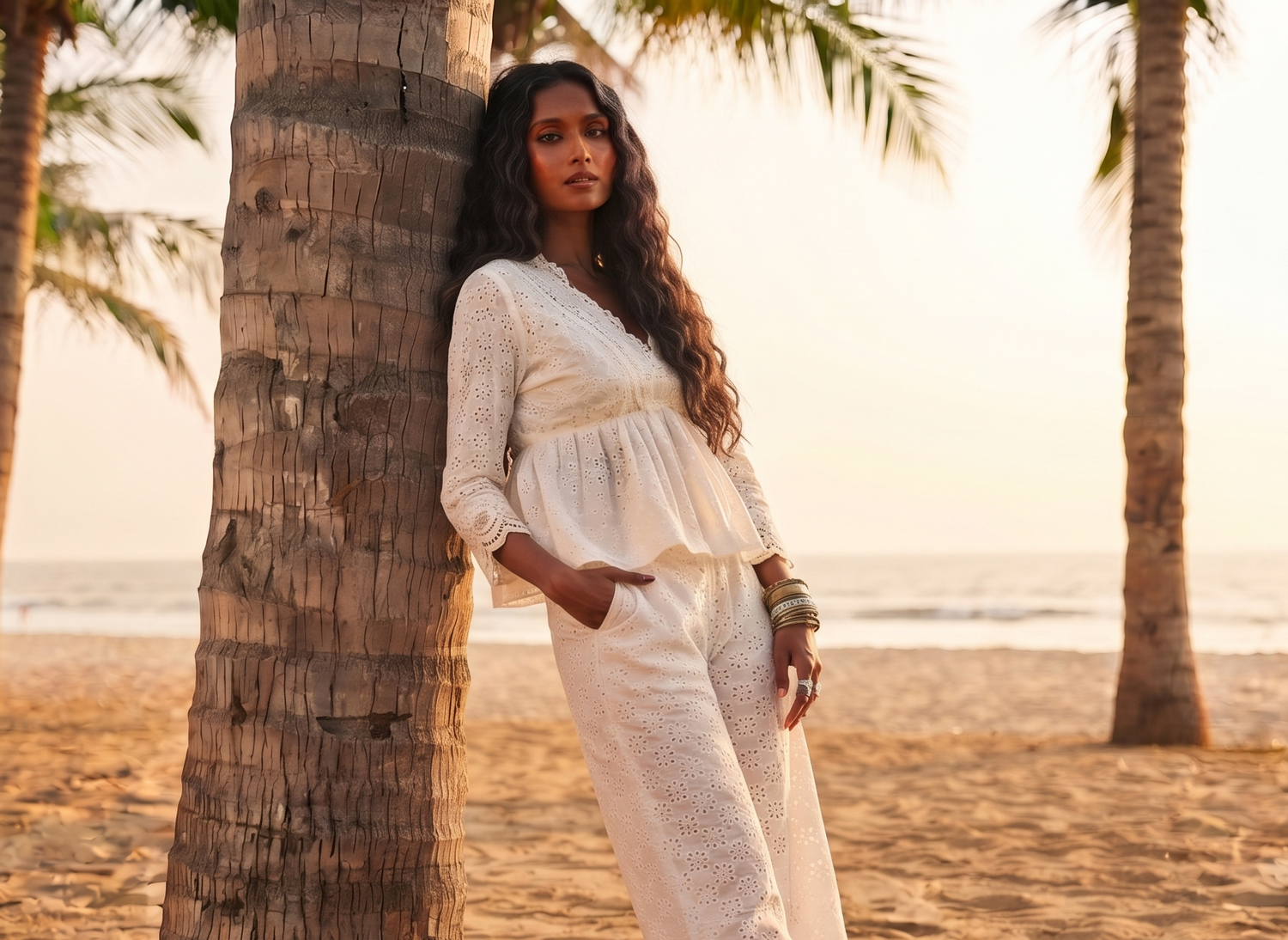 A woman wearing a white embroidered peplum top and matching wide-leg trousers stands on a sandy beach leaning against a palm tree, with the ocean and other palm trees in the background.